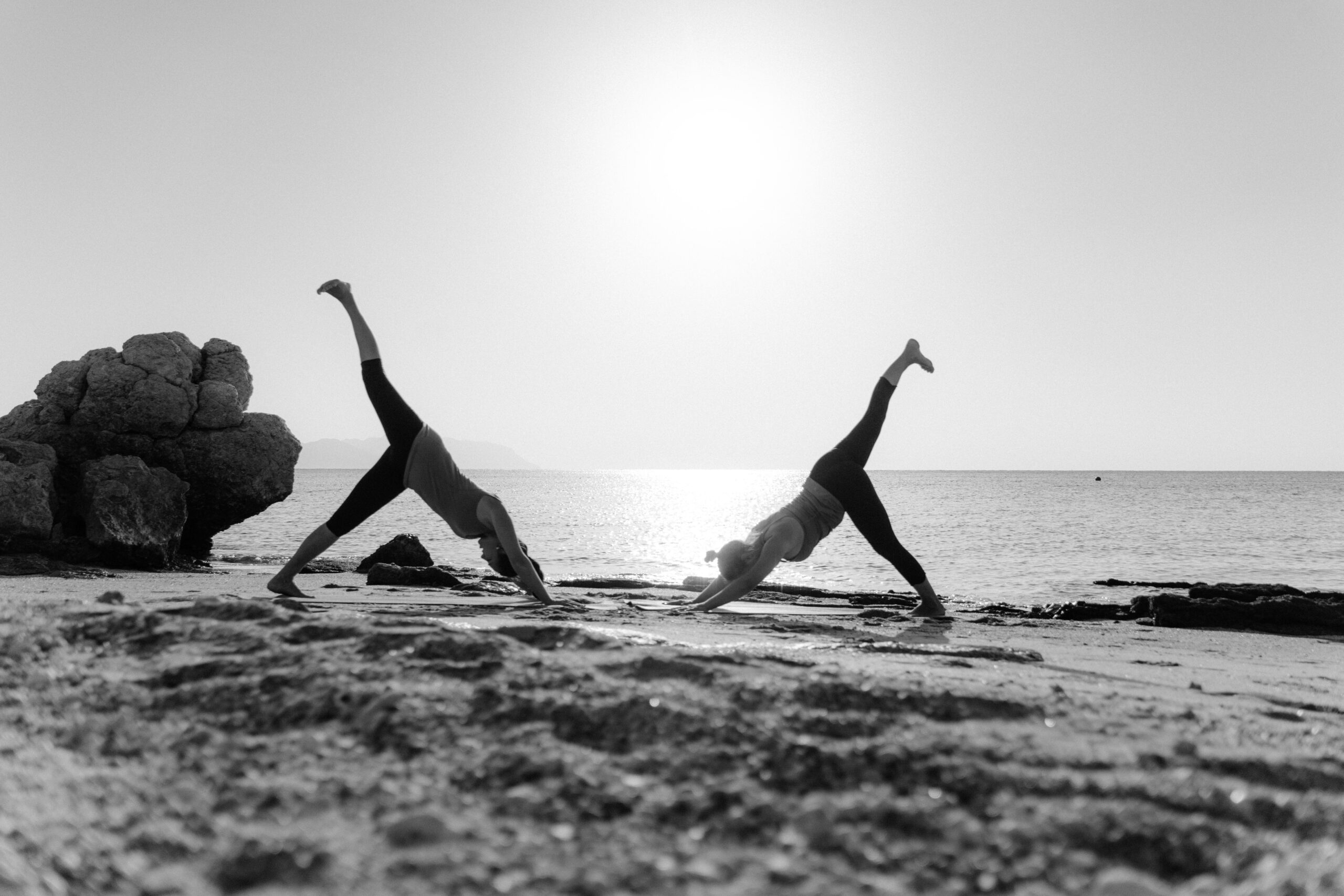 Two women practicing yoga on a beach during sunrise in a serene black and white setting.